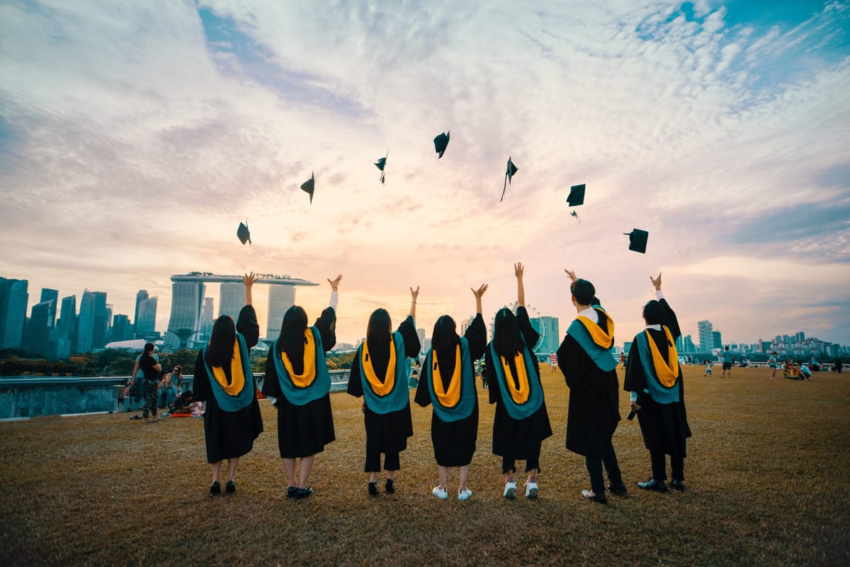 Graduates in gowns and hoods throwing mortarboards into the air in front of a city skyline at sunset