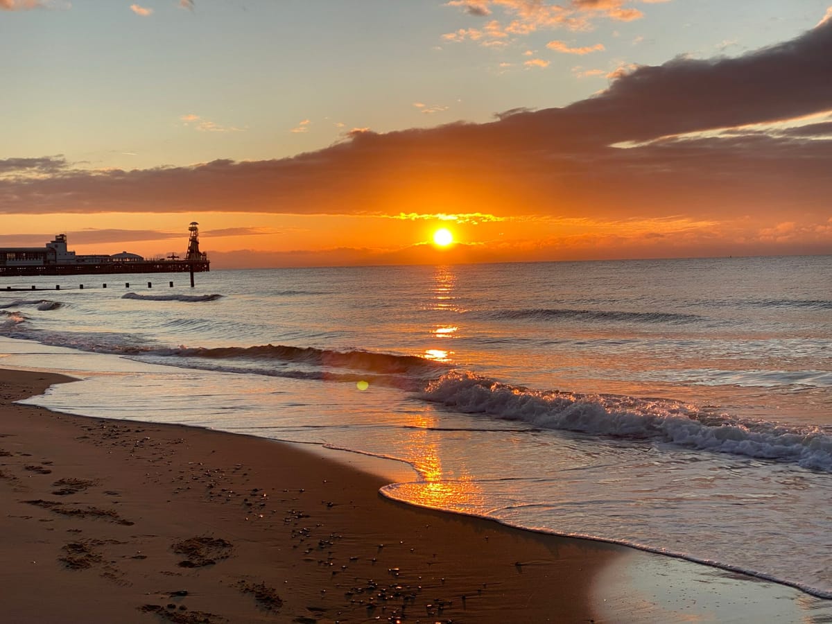 Sun low over the sea from a sandy beach, waves rolling in, a pier in silhouette