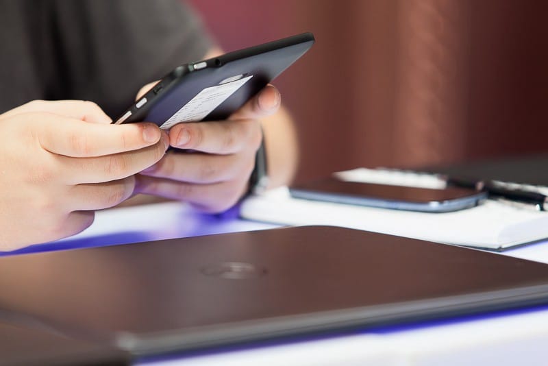 Close-up of hands using a smartphone beside a closed laptop, a second phone and a notebook
