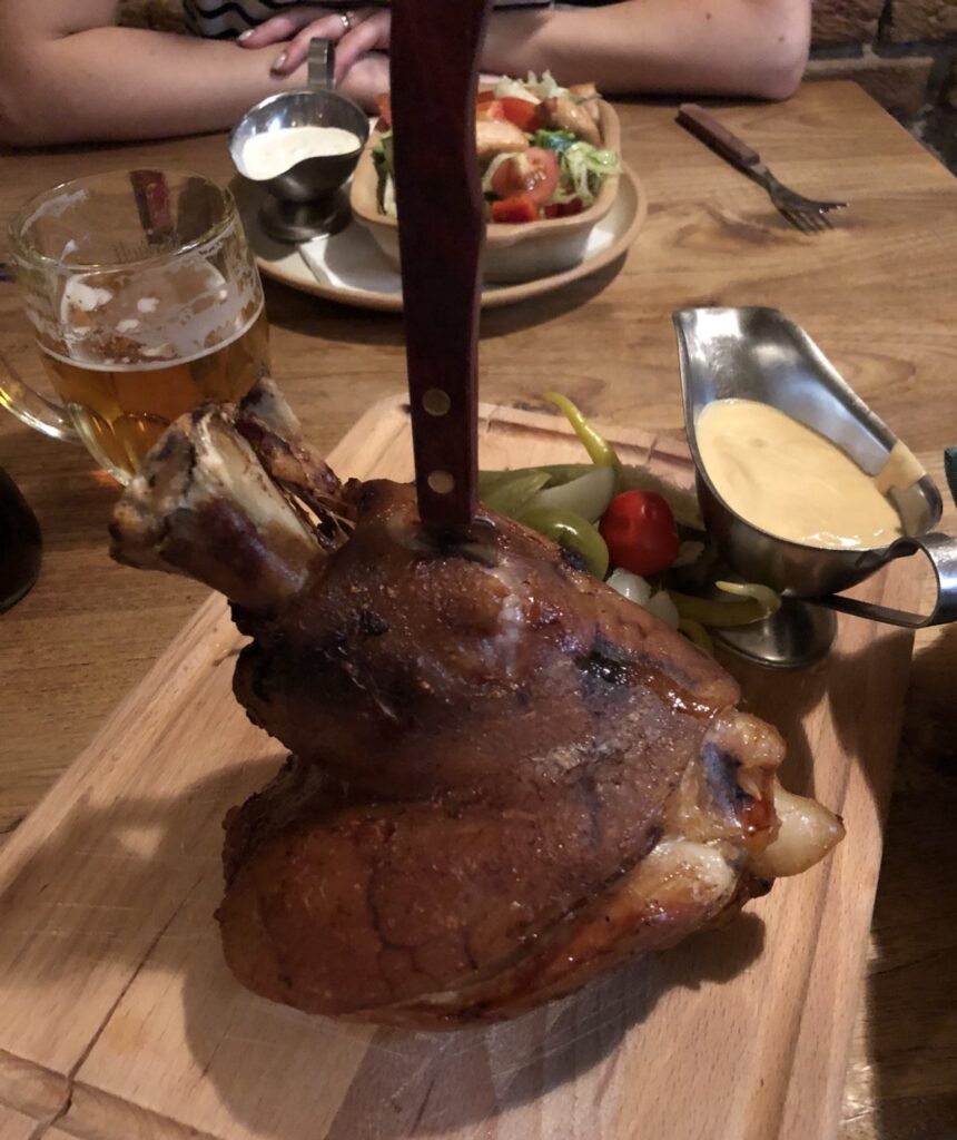Roast pork knuckle with a steak knife stuck in it on a wooden board, with peppers, sauce and beer