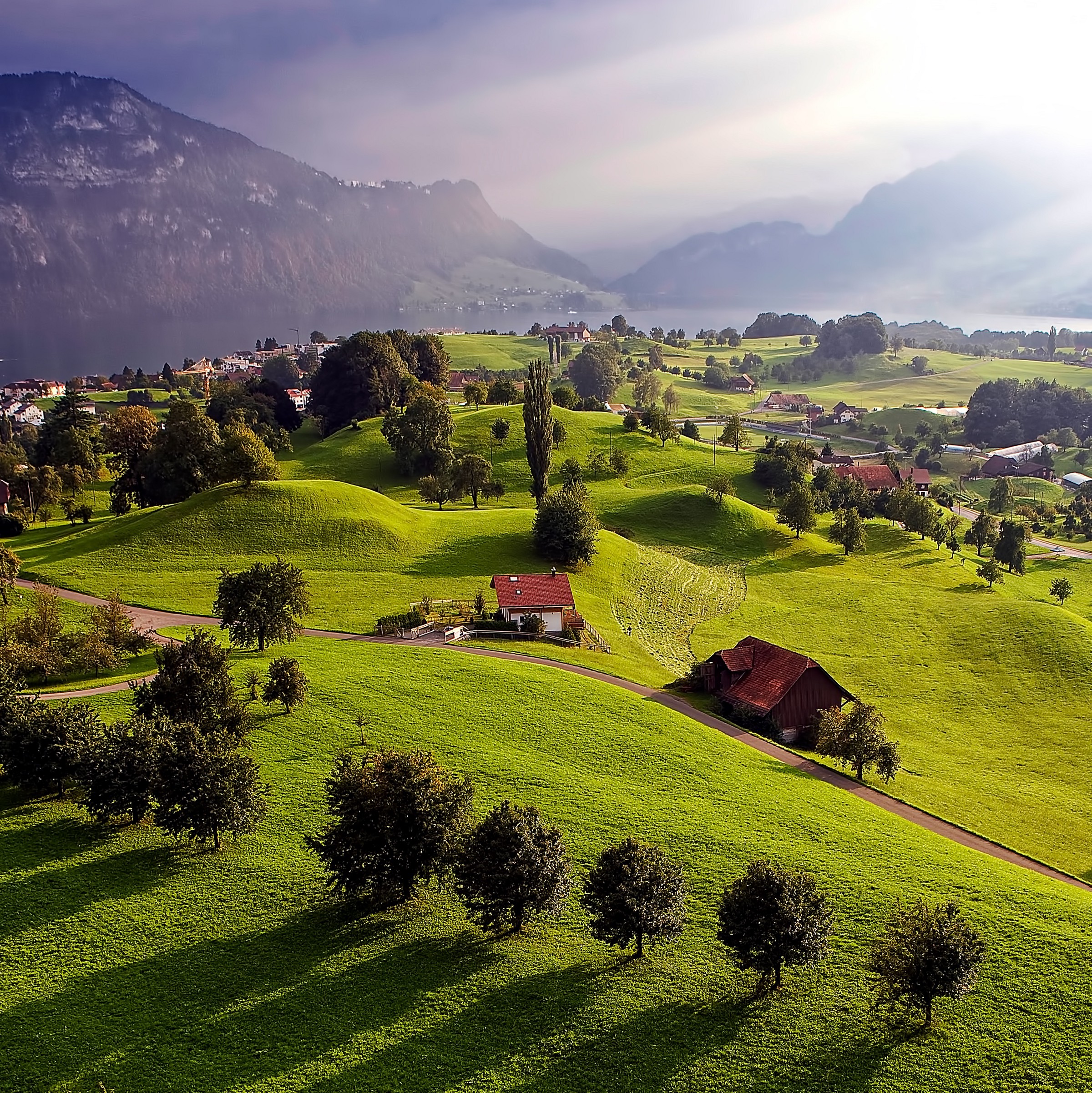 Green hills dotted with farmhouses above a lake, with misty mountains behind