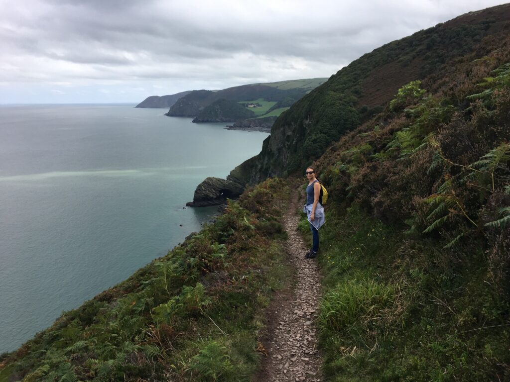 Hiker on a narrow path along steep, fern-covered sea cliffs on the South West Coast Path