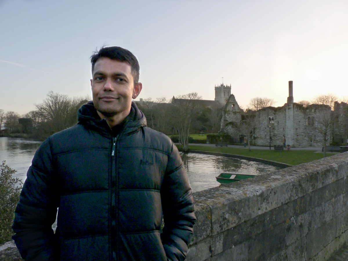 Man in a padded jacket on a stone bridge, a river and priory ruins and church tower behind at dusk