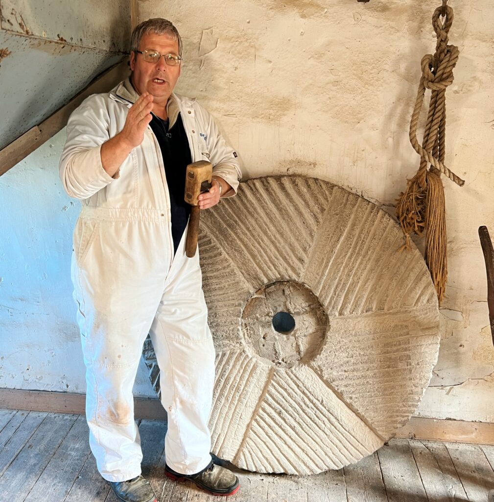 Miller in white overalls holding a mallet beside a large grooved millstone